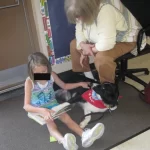 Young girl sits beside a Border Collie lying next to her as she reads a story to thr dog. Dog's handler sit in vhsit next to the dog.