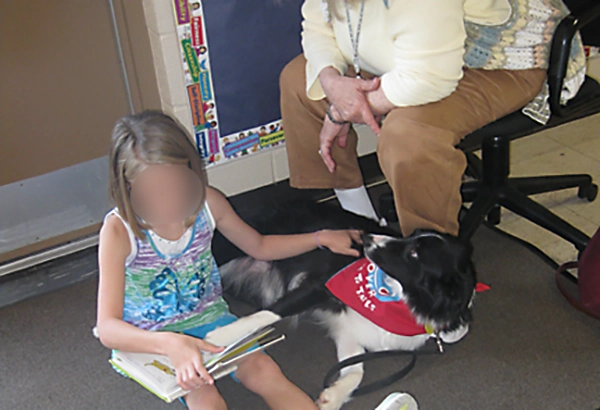 Girl sitting on floor next to Border Collie petting the dog with one hand while holding open a book with the other